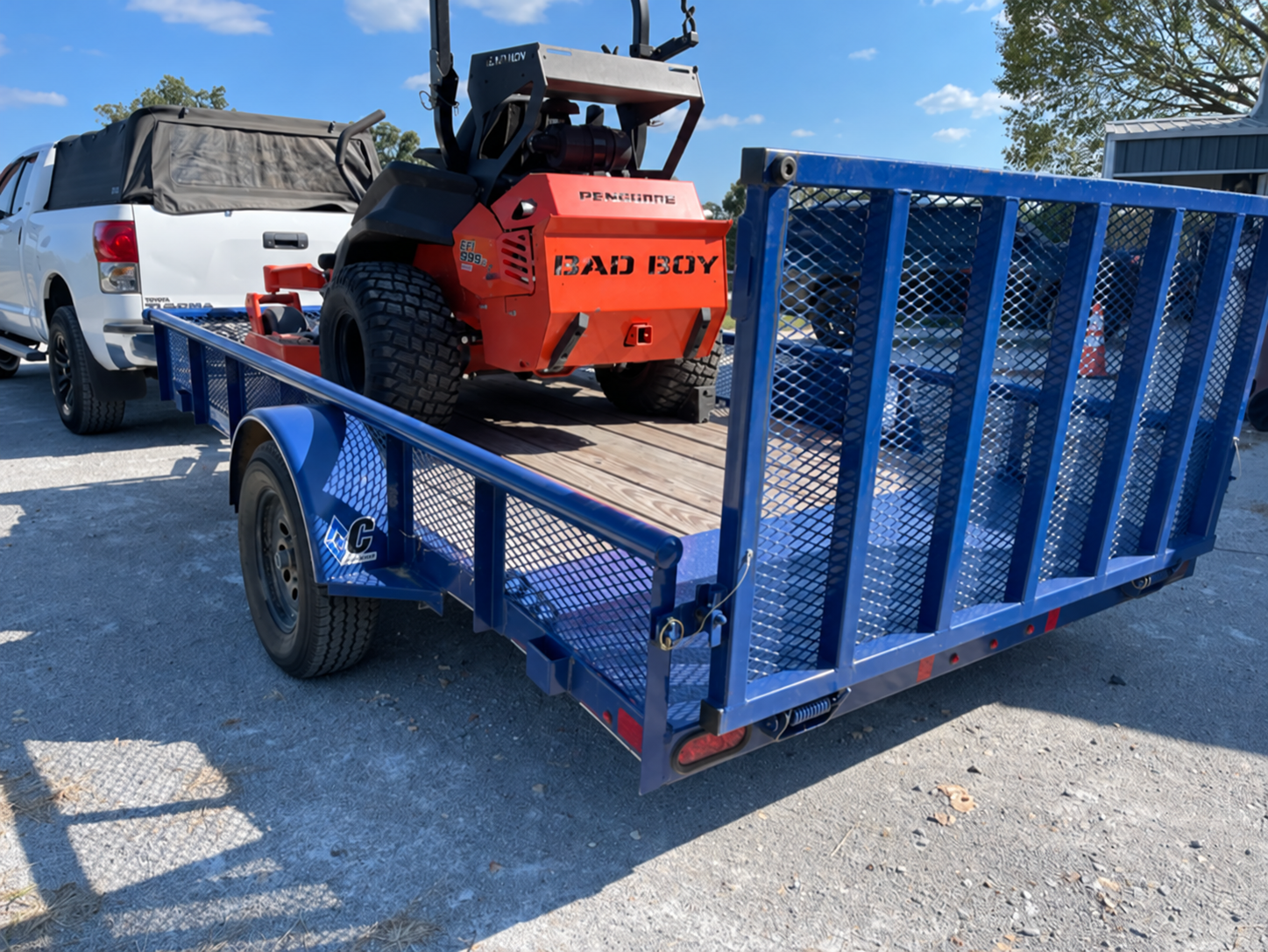 Blue single-axle utility trailer with a rear mesh loading gate, shown carrying a mower